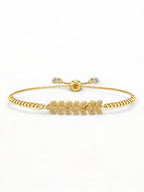 La imagen présente un bracelet élégant et sophistiqué pour femme. Il est composé d'une chaîne en boîte ajustable en acier inoxydable plaqué or 18 carats. Le design met en valeur un charme central en forme de feuille, orné de cristaux brillants CZ (Cubic Zirconia). La chaîne est ajustable, permettant un ajustement parfait à différents poignets, grâce à une perle coulissante qui facilite la personnalisation de la taille. Le bracelet est posé sur une surface élégante, avec un fond gris neutre qui met en avant 